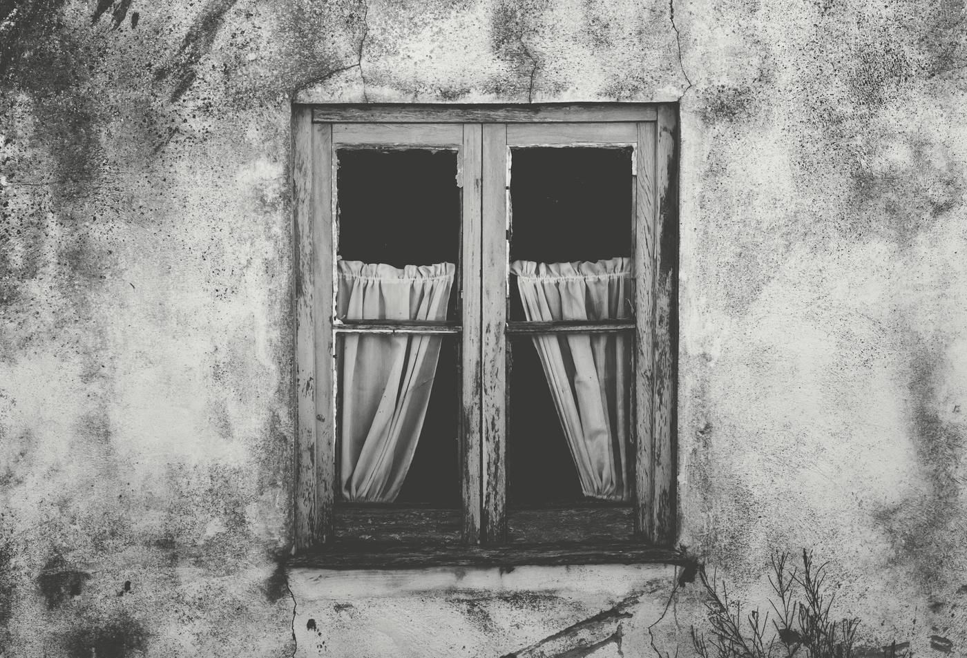 Una ventana vieja con cortinas de encaje en un muro desconchado, en blanco y negro