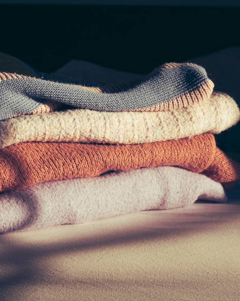 Pila de suéteres en gris, crudo y óxido sobre una superficie en penumbra