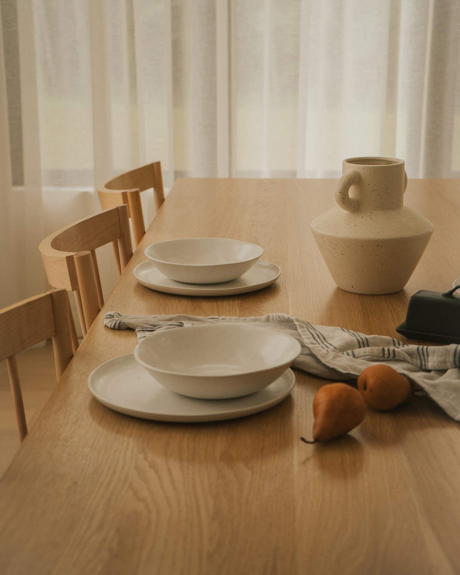 Mesa de madera con vajilla clara, un jarrón y peras junto a una cortina