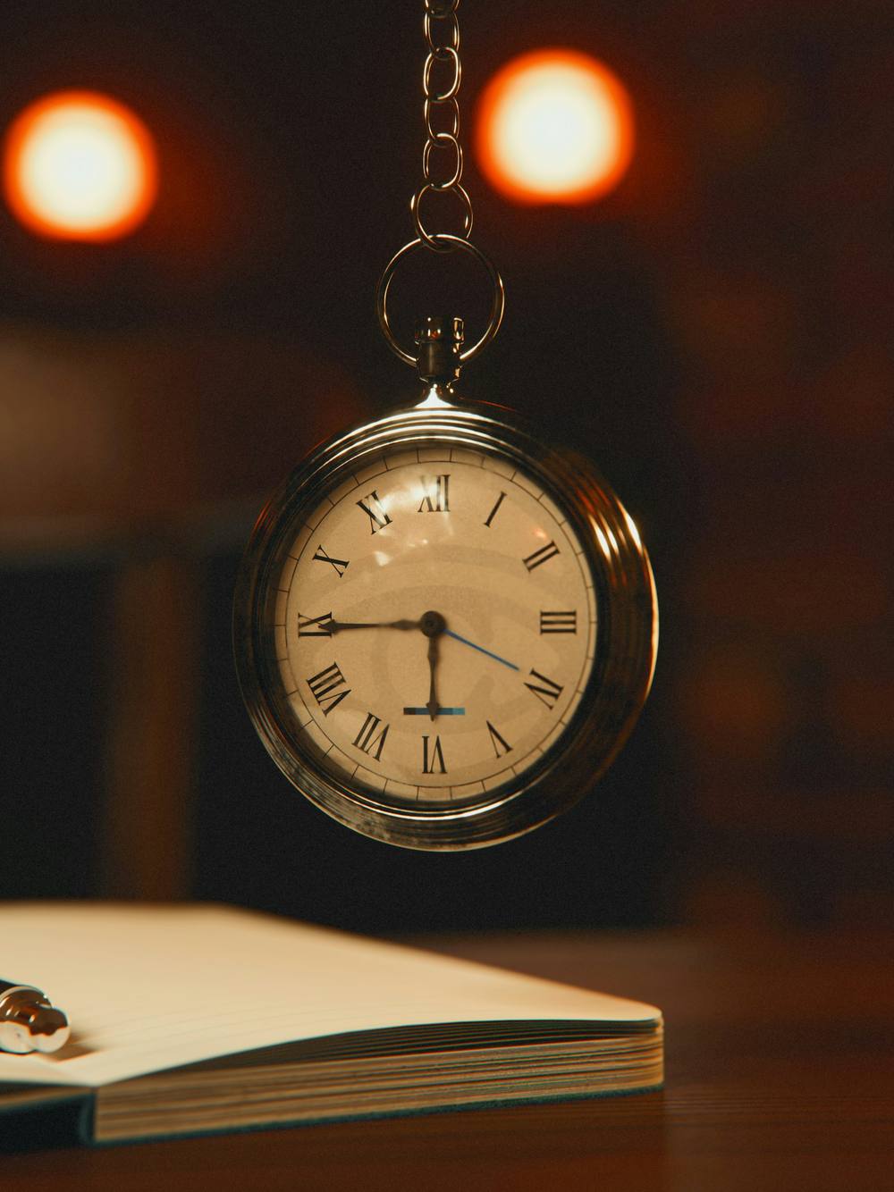 Reloj de bolsillo colgado frente a dos luces cálidas desenfocadas y un cuaderno