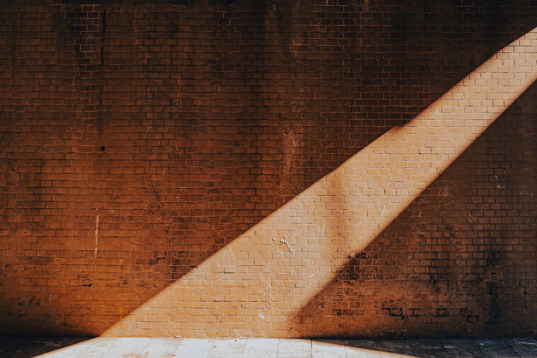 Muro de ladrillo oscuro atravesado por una franja de luz clara que baja en diagonal hasta el suelo