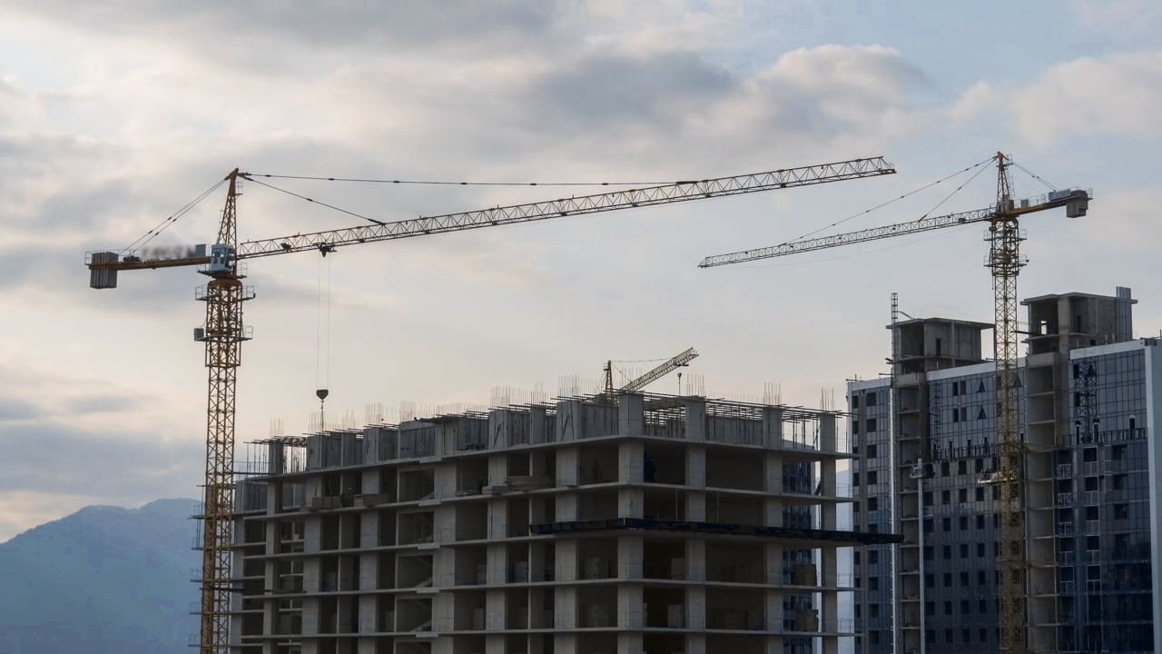 Un edificio de hormigón en obra y otro de vidrio junto a dos grúas torre, bajo un cielo con nubes y montañas al fondo