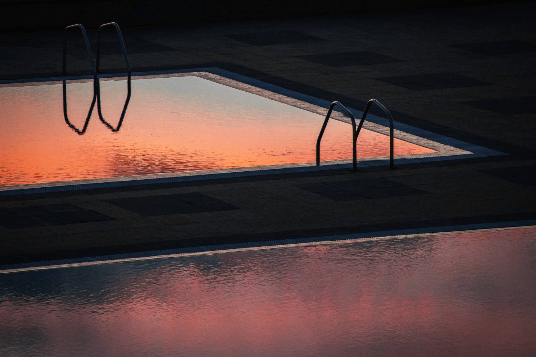 Borde de una piscina oscura que refleja un cielo naranja, con dos barandas de acero