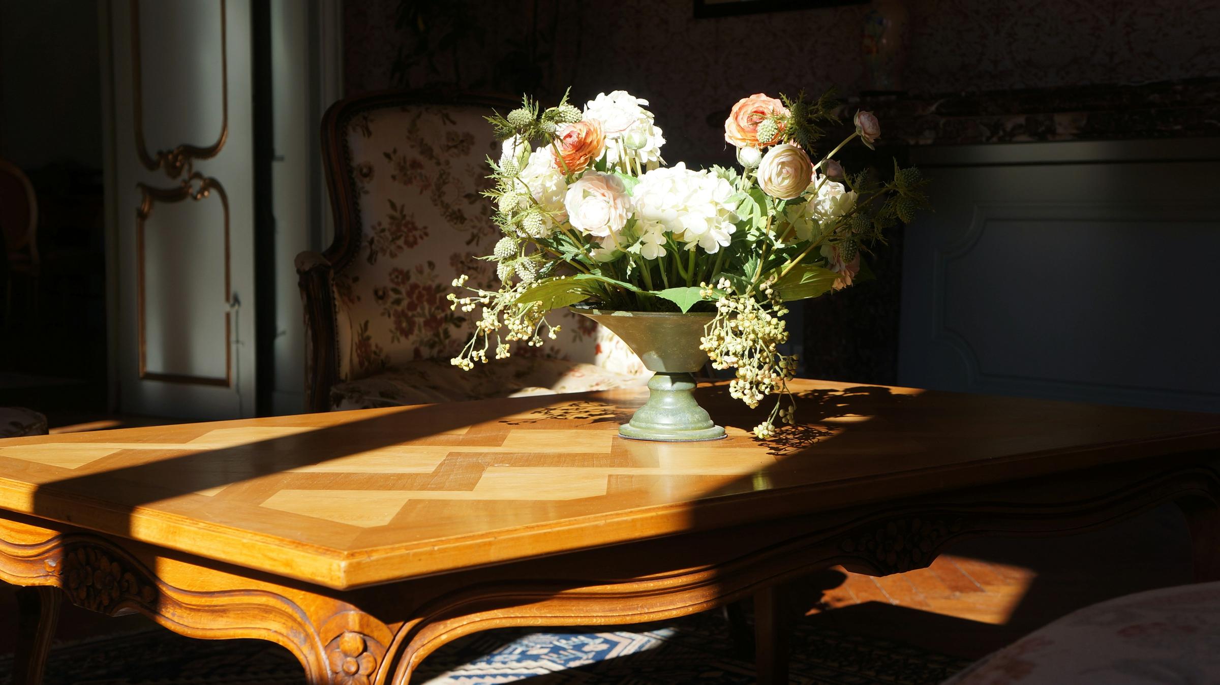 Mesa de madera con marquetería iluminada por el sol, con un arreglo de flores blancas y un sillón al fondo