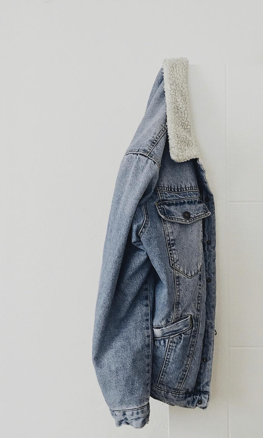 Una chaqueta de mezclilla clara con cuello de borrego colgada de un gancho en una pared blanca