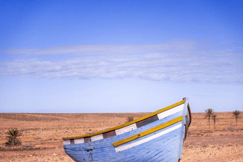 Proa de un bote azul y blanco varado sobre arena naranja, bajo un cielo azul