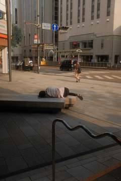 Una persona duerme sobre una banca de piedra en una plaza vacía, con un peatón al fondo