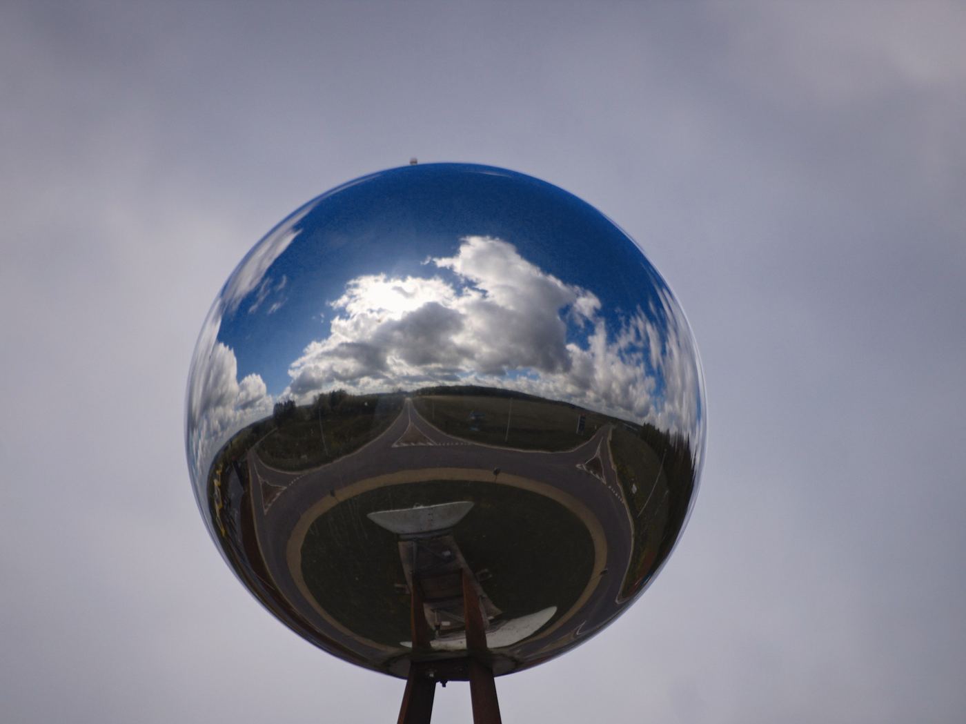 Esfera de acero pulido sobre un soporte de metal que refleja el cielo nublado y el paisaje
