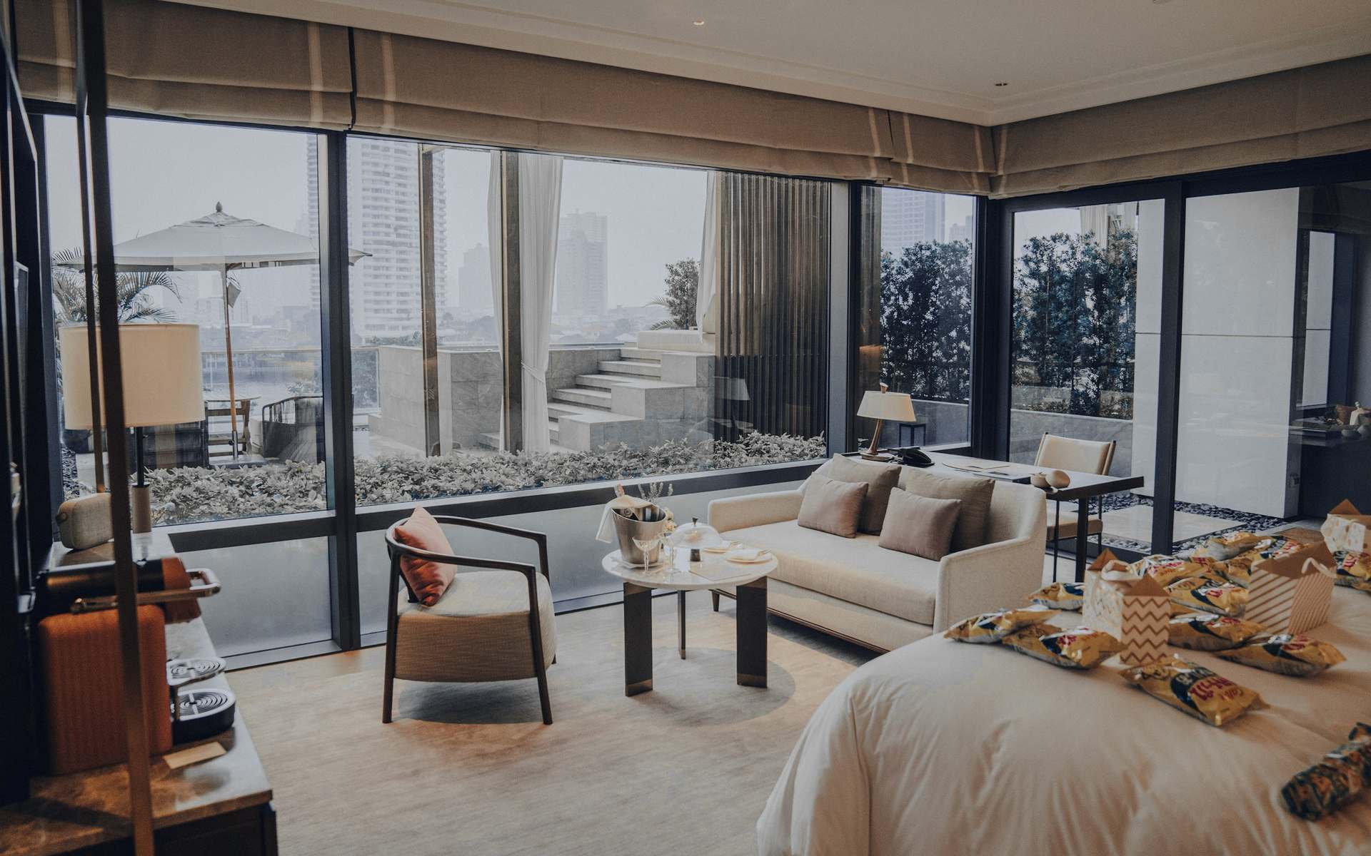Salón con grandes ventanales, sofás claros y una cama deshecha al fondo