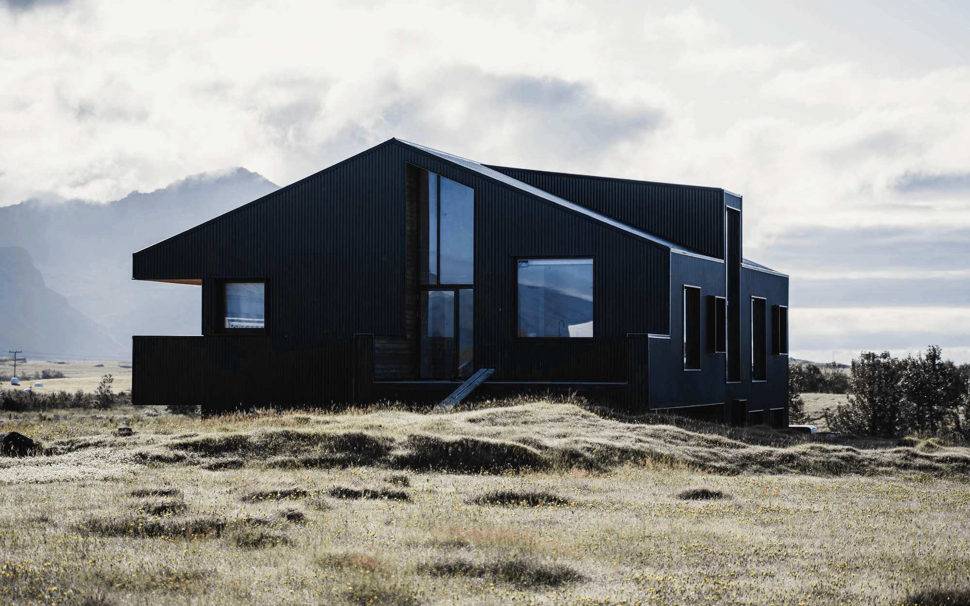 Casa negra de líneas quebradas en una pradera de altura, bajo nubes y montañas