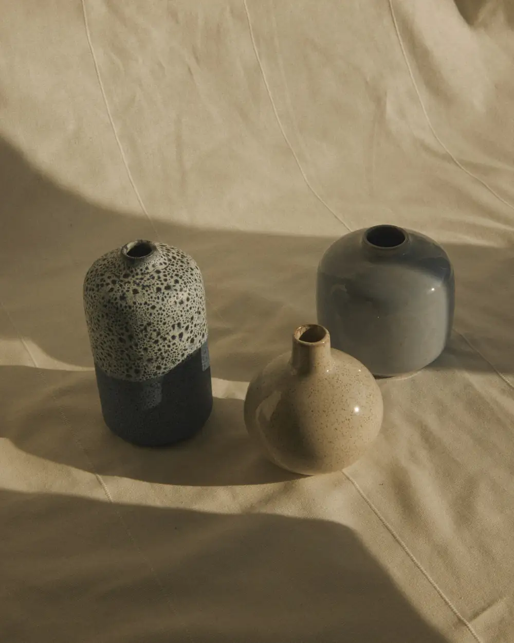 Tres botellas esmaltadas, una moteada y otra gris, sobre una tela de lino arrugada