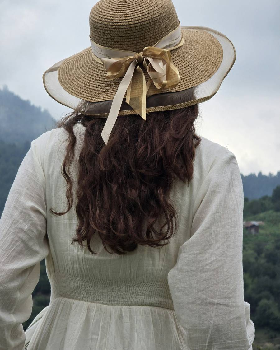 Persona de espaldas con vestido blanco y sombrero de paja frente a las montañas
