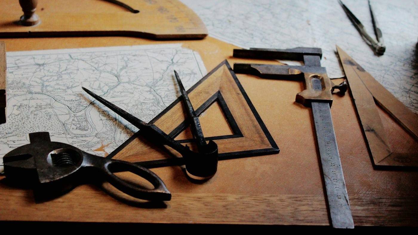 Escuadra, compás y reglas antiguas sobre una mesa de madera, con mapas de fondo