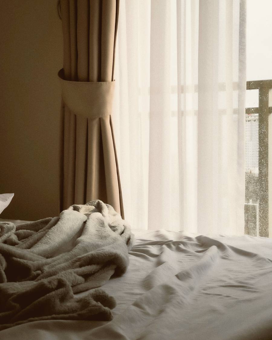 Una cama deshecha junto a un ventanal, con una cortina de lino recogida y un velo claro detrás