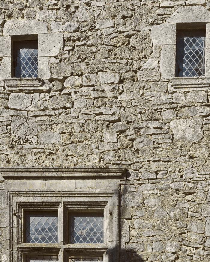 Muro de piedra con dos ventanitas de vidrios en rombos y, más abajo, una ventana mayor con marco de piedra