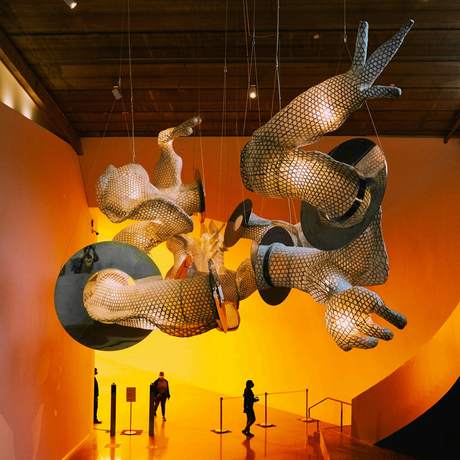 Escultura metálica suspendida en una sala anaranjada, con dos siluetas mirándola