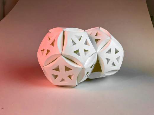 Escultura de papel blanco con huecos en forma de estrella, iluminada en rosado y celeste