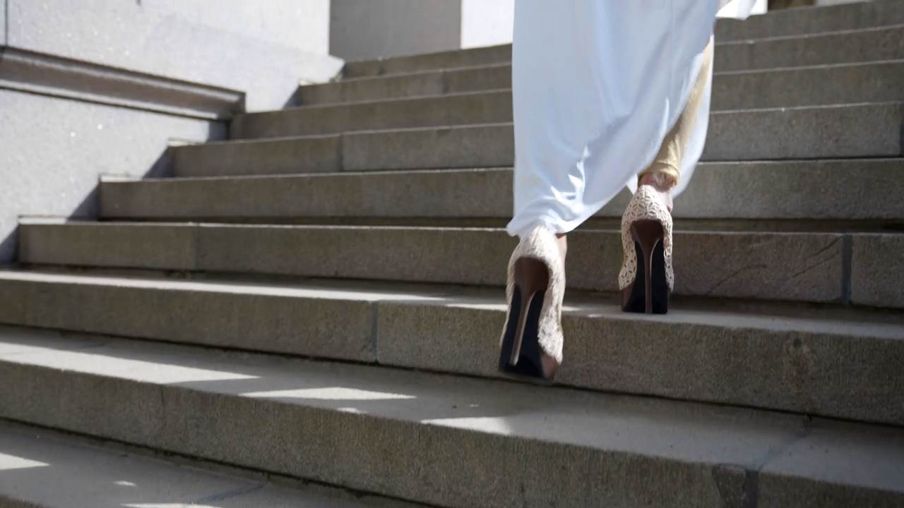 Una persona con vestido blanco sube una escalinata de piedra; solo se ven las piernas y los zapatos