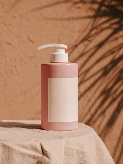 Botella rosada con dosificador sobre un paño de lino, con sombras de hojas en una pared terracota