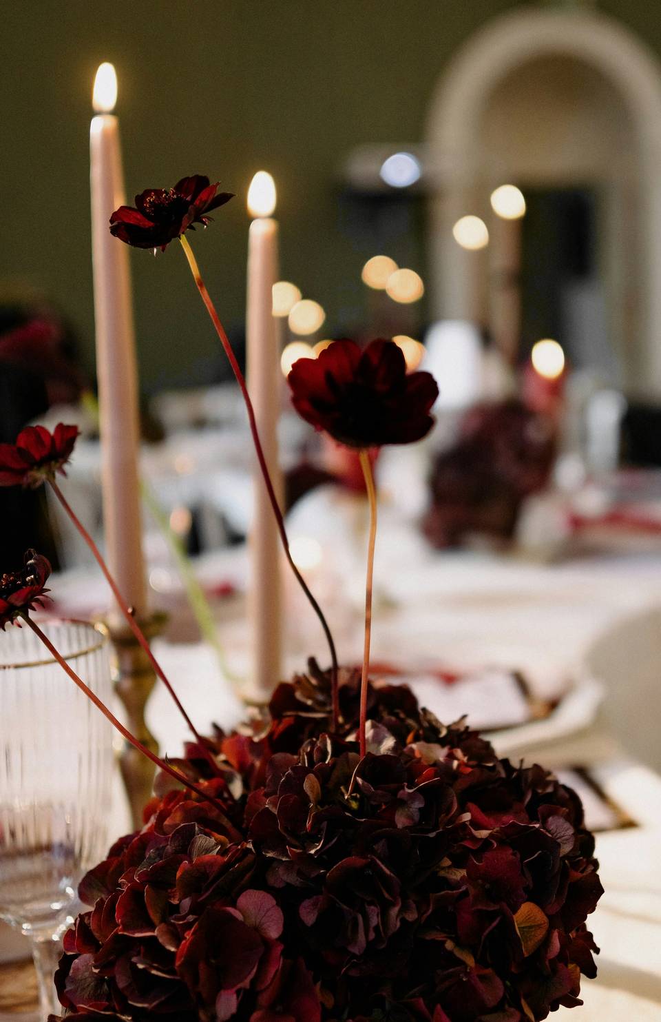 Velas encendidas y hortensias secas en un centro de mesa, con copas al fondo
