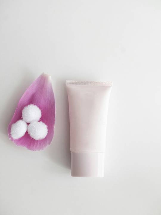 Tubo rosado de crema junto a un pétalo y bolitas de algodón, sobre un fondo blanco