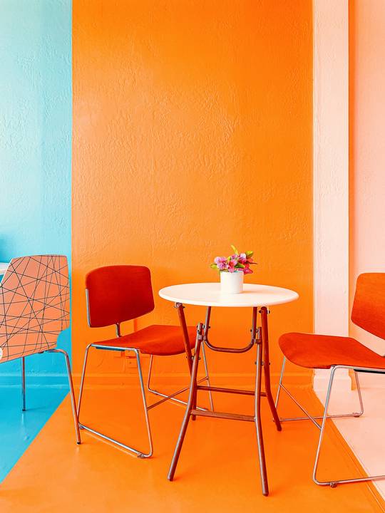 Rincón naranja con una mesa blanca y sillas rojas sobre un piso azul