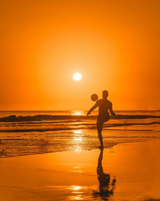Silueta de una persona que patea una pelota junto al mar, con el sol detrás