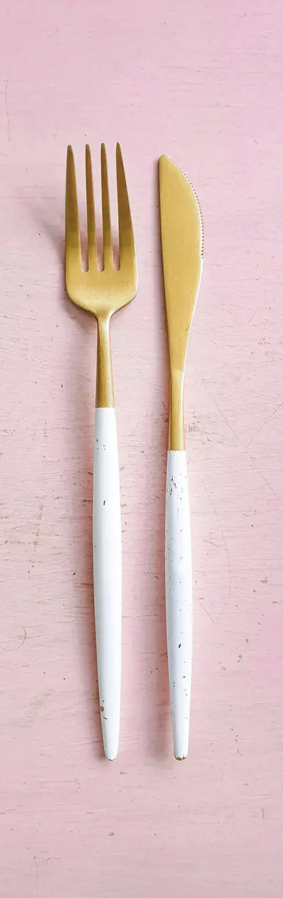Tenedor y cuchillo dorados con mango blanco sobre un fondo rosado