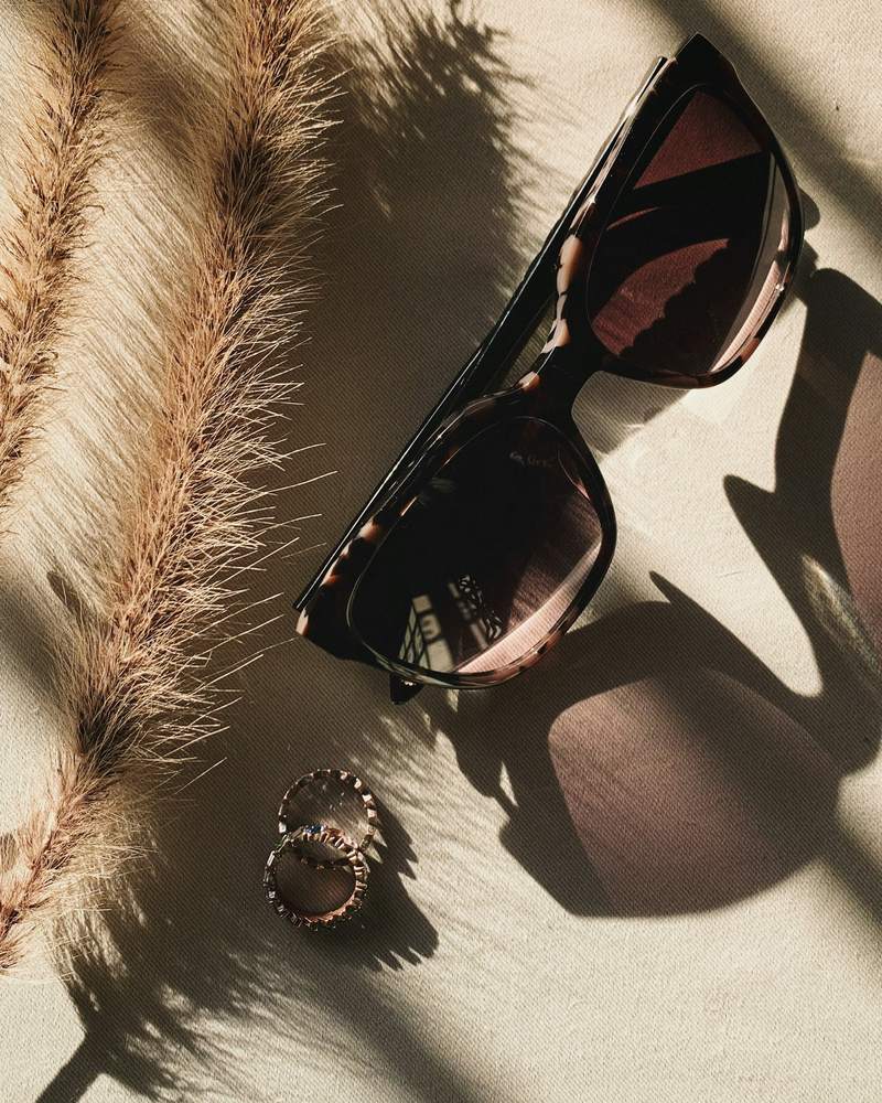 Lentes de sol junto a plumas de pasto seco y sus sombras sobre una superficie arenosa