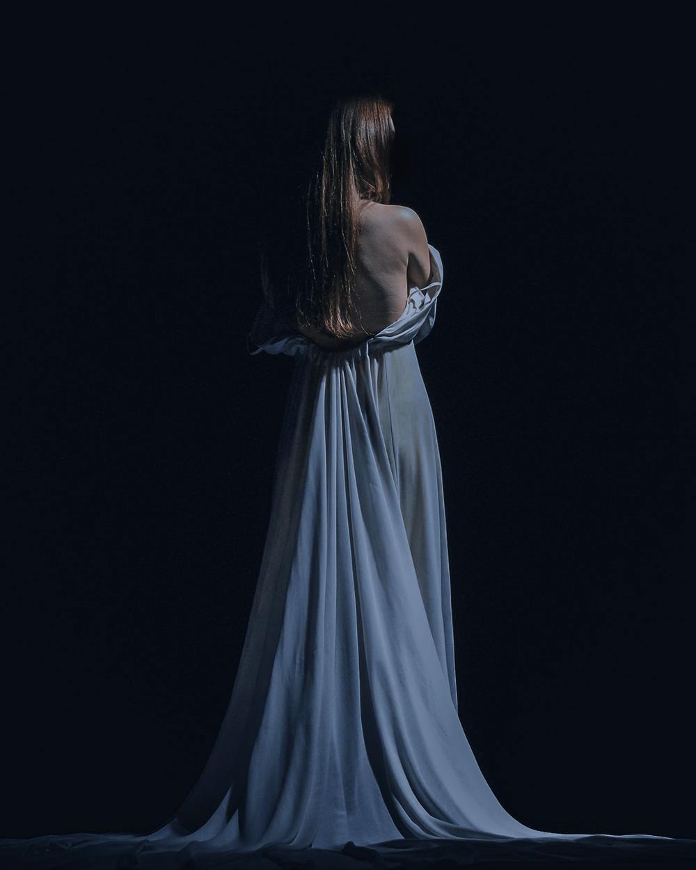 Una mujer de espaldas con un vestido largo azul pálido sobre un fondo oscuro