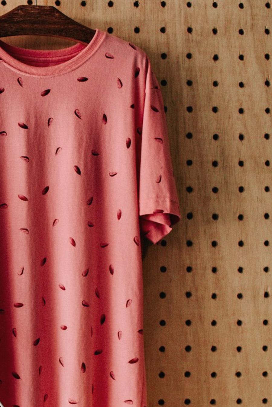 Una camiseta rosa con un estampado de semillas oscuras colgada de una percha de madera sobre un tablero perforado