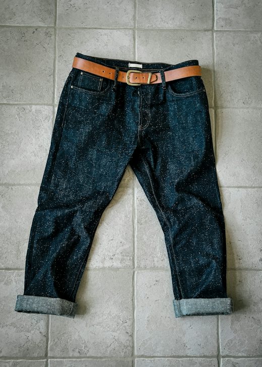 Pantalón de mezclilla oscura extendido sobre baldosas, con un cinturón de cuero