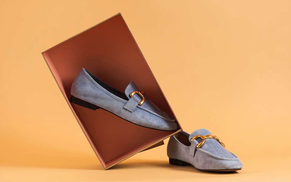 Mocasines de ante gris azulado, uno dentro de una caja color terracota sobre un fondo naranja claro