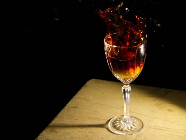 Copa de licor ámbar con un chorro que salpica, sobre una mesa de madera iluminada contra un fondo negro