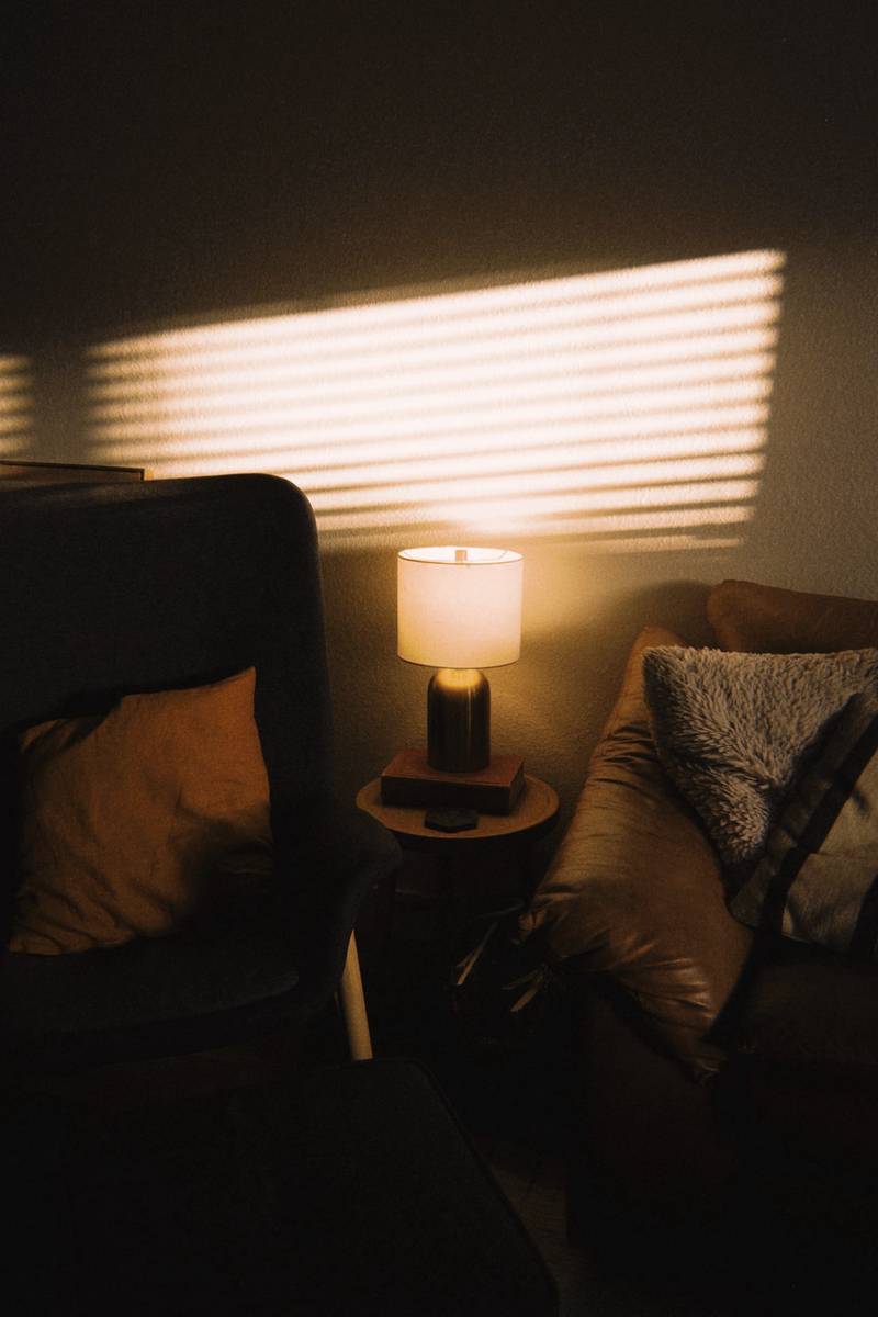 Sillón oscuro con una lámpara encendida y franjas de luz en la pared