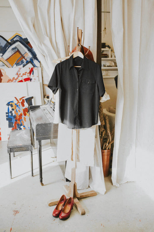 Camisa negra de manga corta colgada de una percha, con unos zapatos rojos al pie y cortinas claras