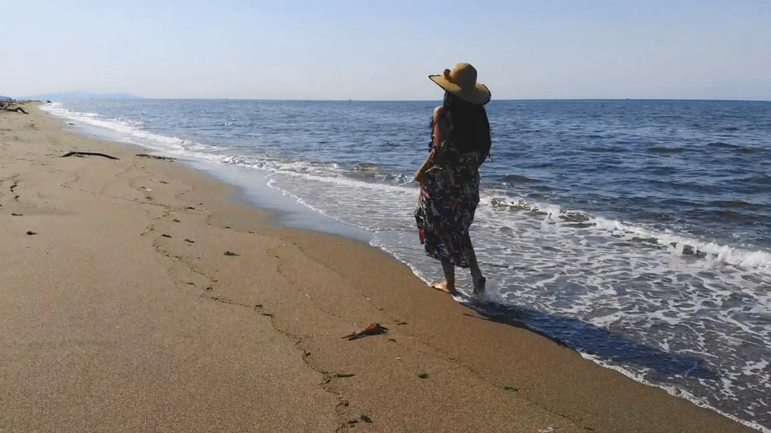 Mujer de espaldas con sombrero de paja y vestido estampado, caminando por la orilla de una playa