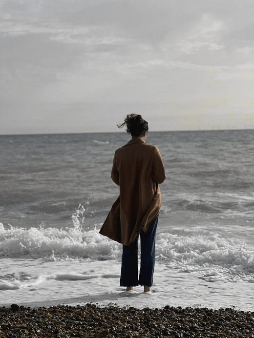 Mujer de espaldas con abrigo marrón, de pie frente al mar con las olas rompiendo en la orilla