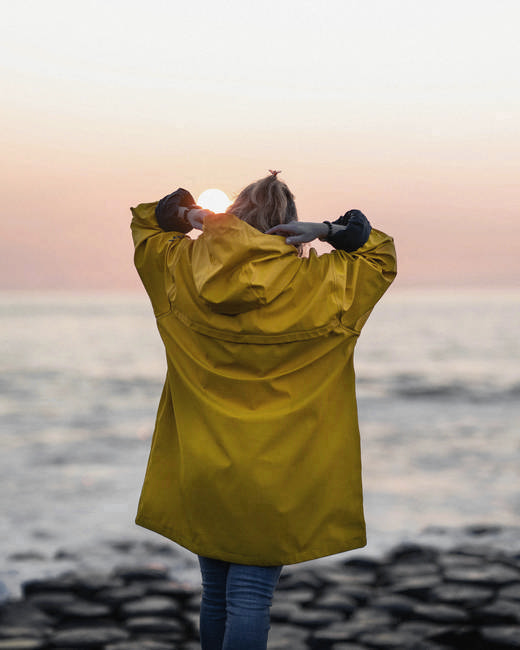 Persona de espaldas con impermeable amarillo, con las manos en la cabeza frente al mar y el sol bajo