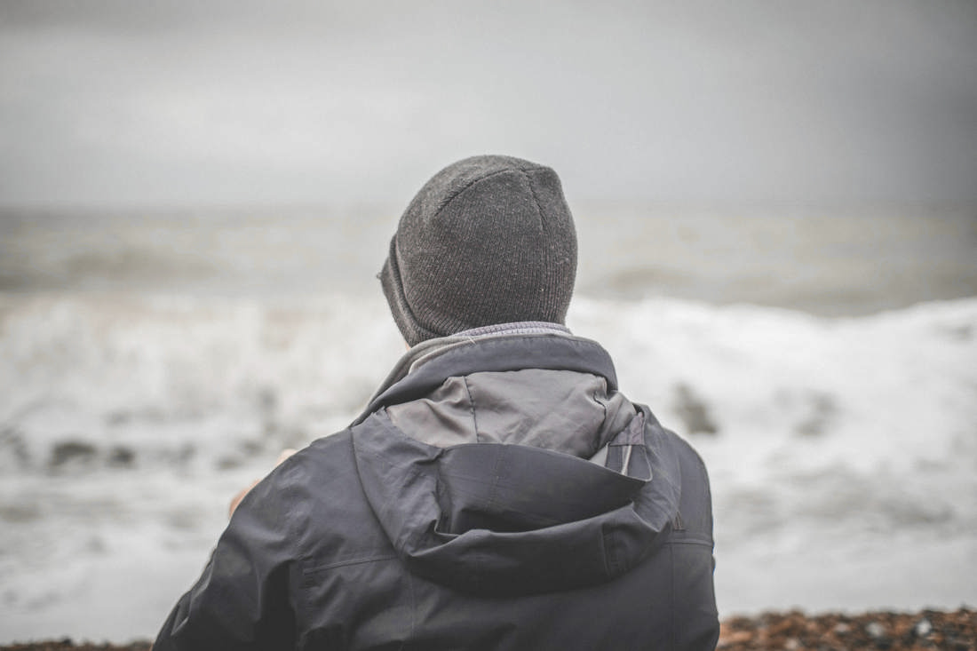 Hombre de espaldas con gorro y chaqueta gris, frente a las olas