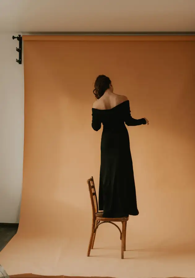 Figura de espaldas con vestido negro sobre una silla, ante un fondo naranja